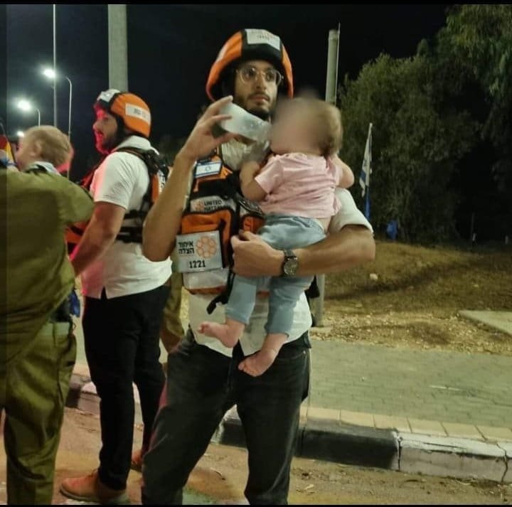 A man in glasses wearing an orange helmet and a first responder vest carries a baby in his arms at night, with blurred figures in the background suggesting a scene of emergency response.