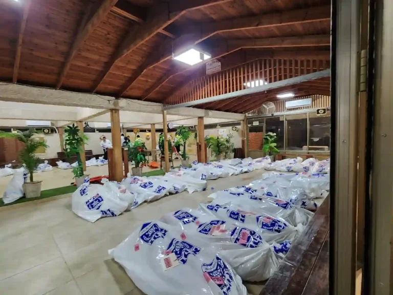 A large room with body bags on the floor, ceiling fans, and a wooden mezzanine with people in the background.