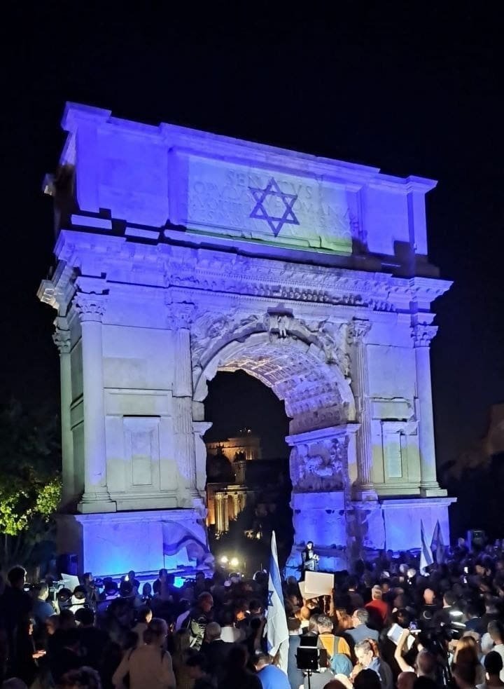 The Victory Gate in Italy illuminated with the colors of the Israeli flag at night, with a crowd gathered in front.