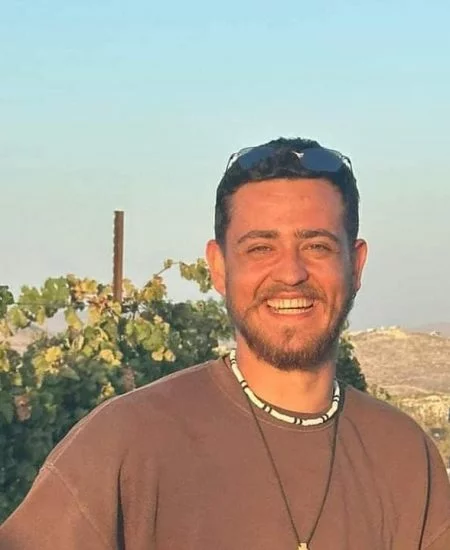 A young man with a kind smile, outdoors with a clear sky and greenery in the background.