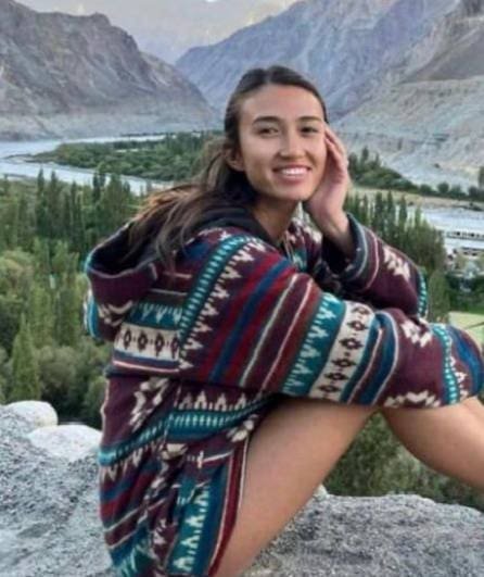 A smiling young woman named Noa sitting on a rock with a scenic valley and river in the background.