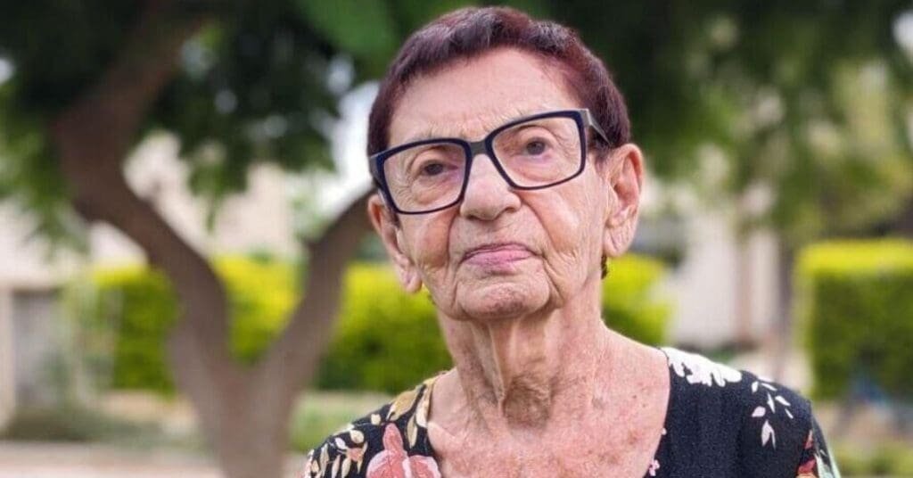 Portrait of an elderly woman with a dignified expression, wearing glasses, with greenery in the background.