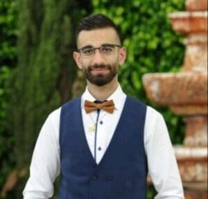 A man with glasses, wearing a bow tie and vest, smiles for a portrait in an outdoor setting.