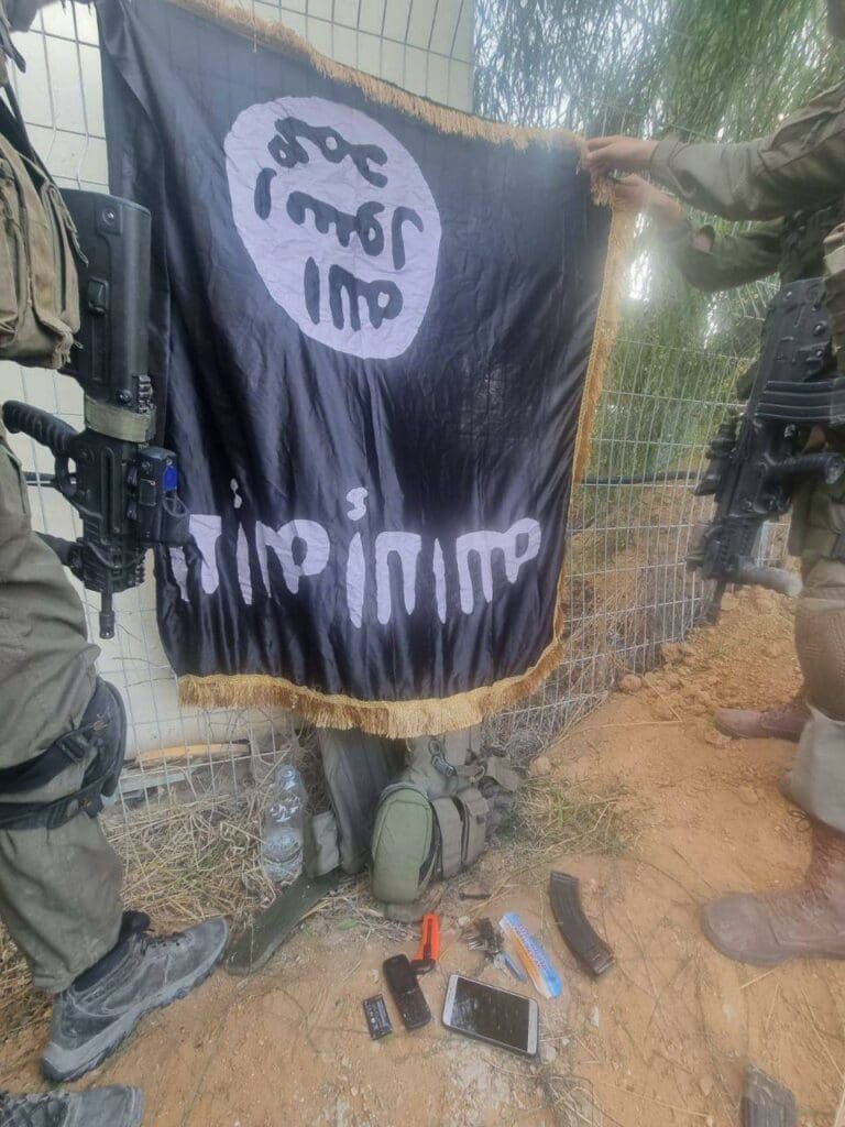 Israeli soldiers hold up a black flag with white Arabic text, identified as an ISIS flag, found among Hamas terrorists' equipment at Kibbutz Sufa.