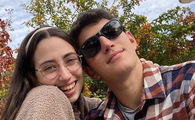 A young couple embracing, smiling at the camera, with autumn foliage in the background.