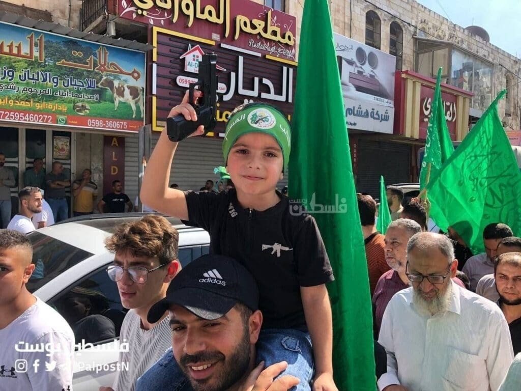 A young child, in a black shirt and green Hamas cap, is raising a toy gun in the air while being carried on someone's shoulders at a public demonstration.