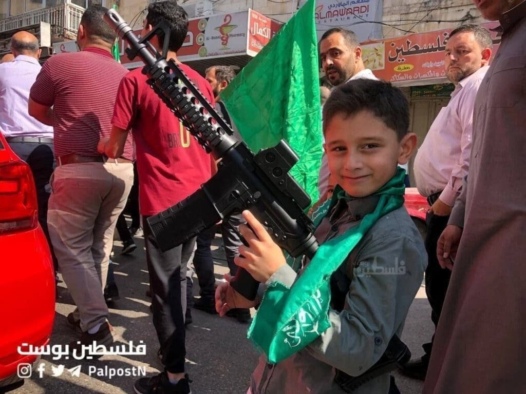 A boy, smiling, holding a large toy gun, with a green Hamas scarf around his neck, at a support Hamas rally.