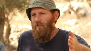 A man with a full beard and wearing a bucket hat speaks with earnest expression in an outdoor setting.