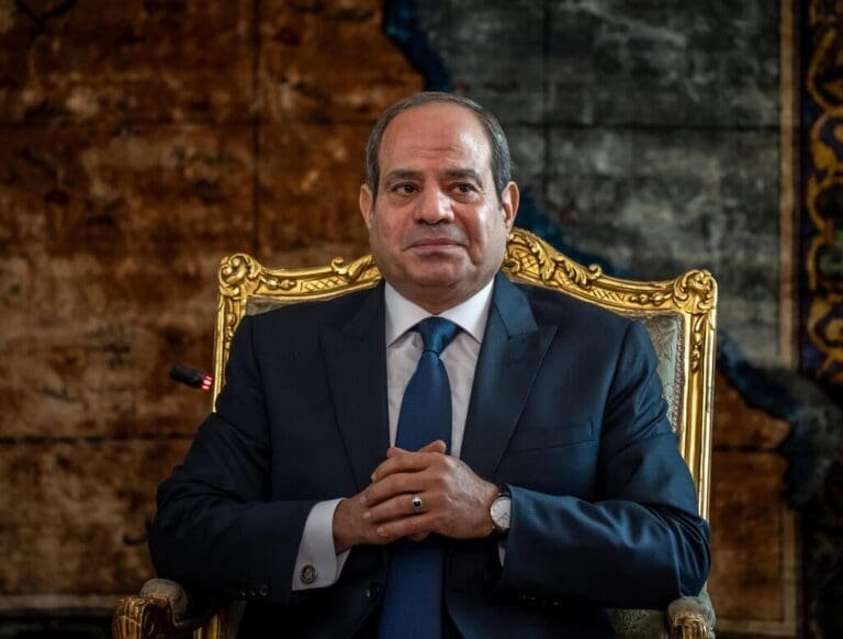 A portrait of a man in a dark suit sitting on an ornate gold chair, with a contemplative expression and hands clasped together.