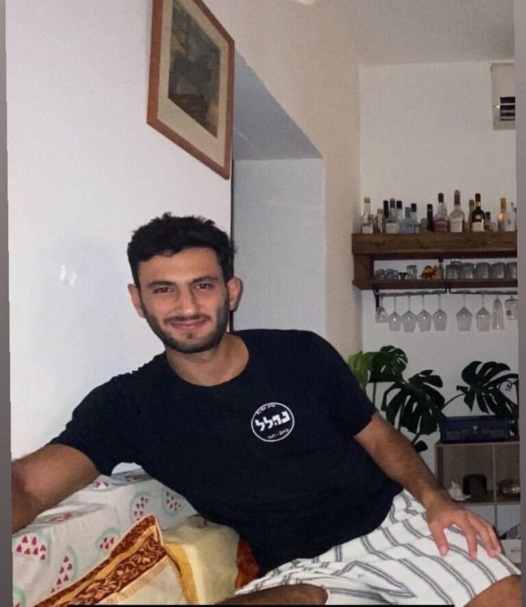 A smiling young man sitting on a sofa, wearing a black t-shirt with a logo.