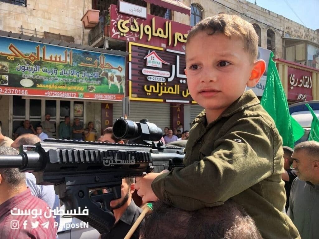 A young child, dressed in olive green attire, is perched on someone's shoulders at a public gathering, holding onto a mock-up of a military-style rifle.