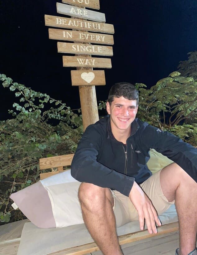 A smiling young man sitting under a sign with an affectionate message, captured in a candid moment of joy.