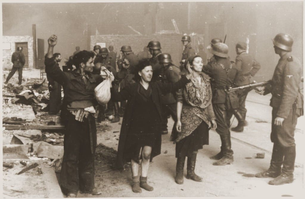 Captured Jewish resistance fighters raise their hands in surrender as they are surrounded by armed SS troops during the Warsaw Ghetto Uprising. The scene depicts the aftermath of the intense struggle within the ghetto, with bodies and destruction visible in the background.